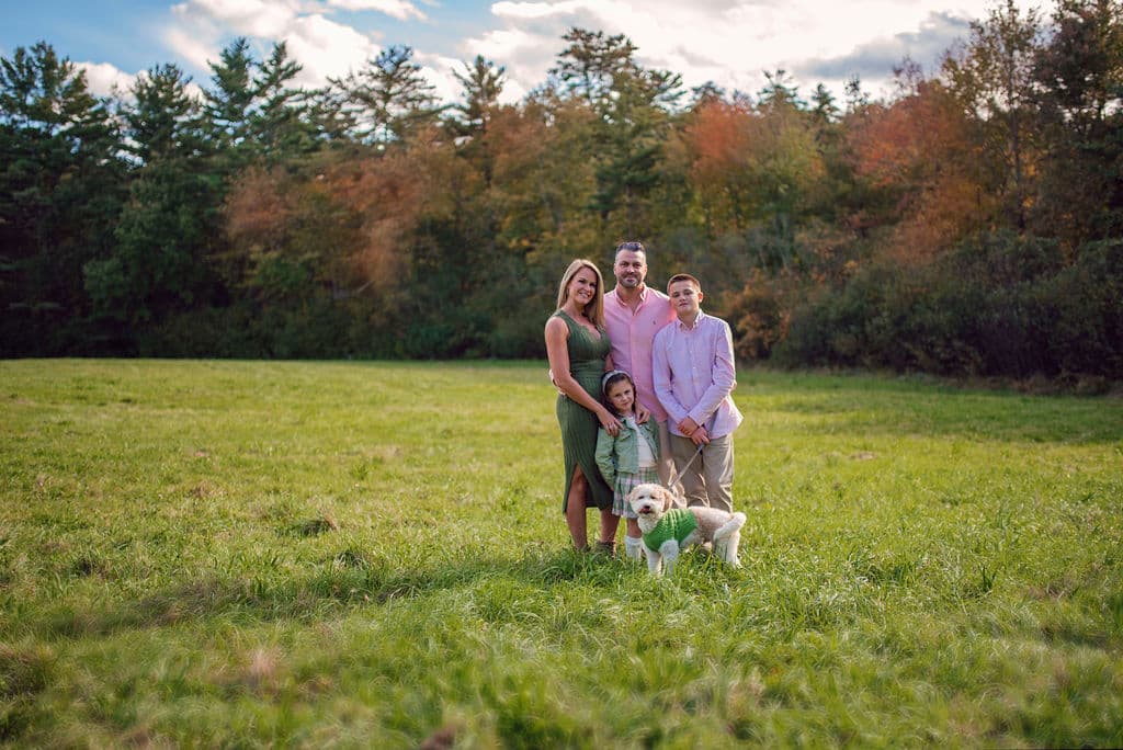 Family with their dog in a wide green field framed by fall trees