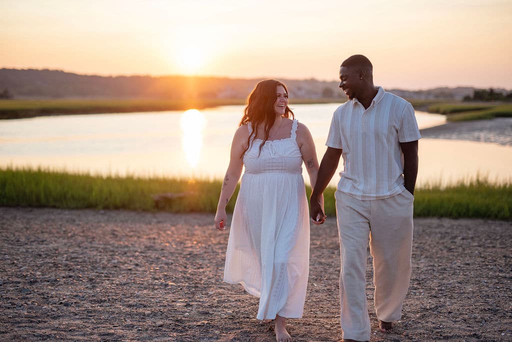 Expecting couple walking hand in hand by the water at sunset