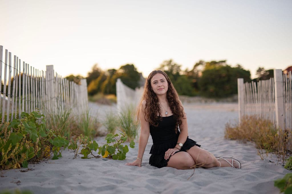 Senior girl sitting in the sand beside a weathered beach fence