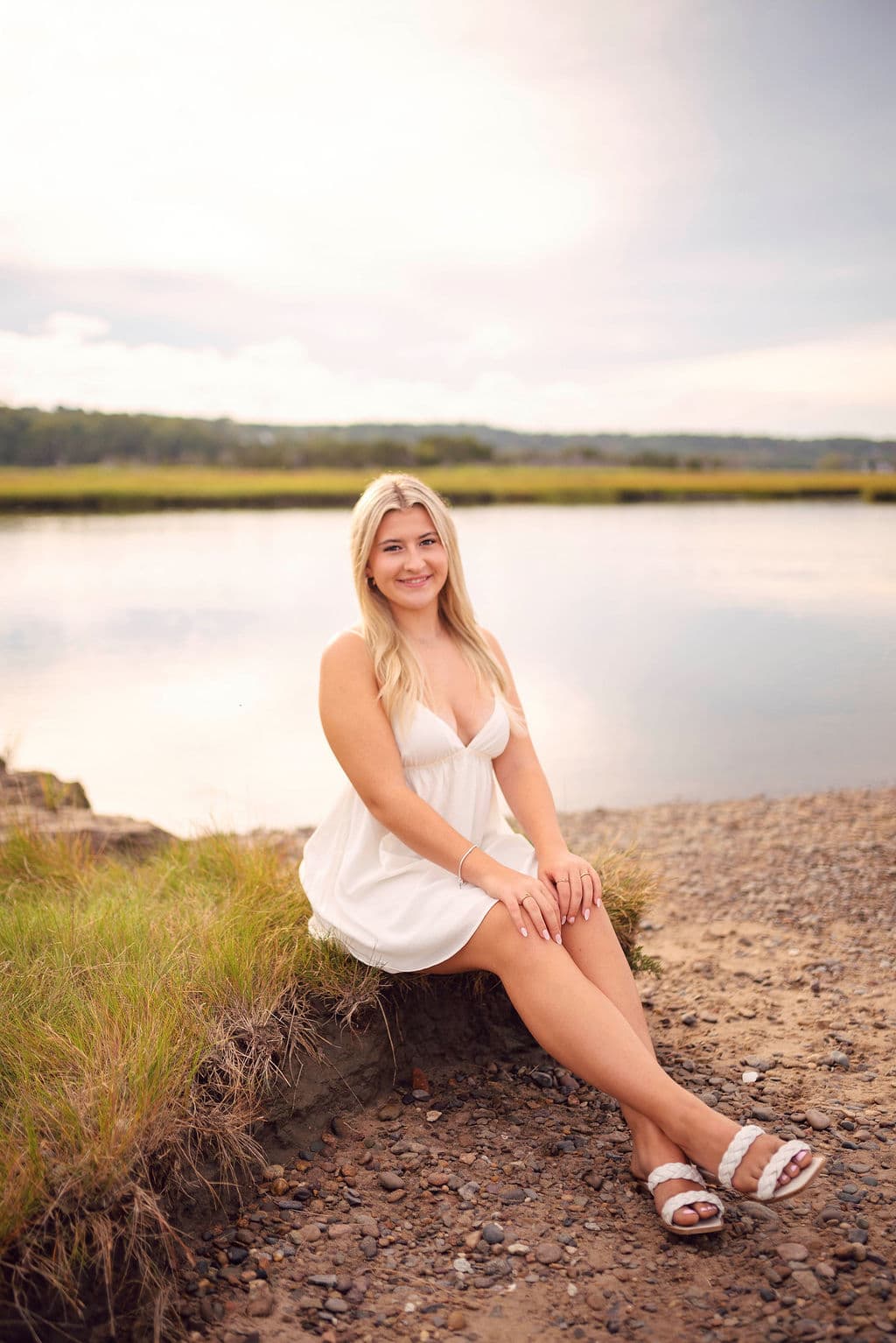 Senior girl in a white dress sitting on a grassy pond shore